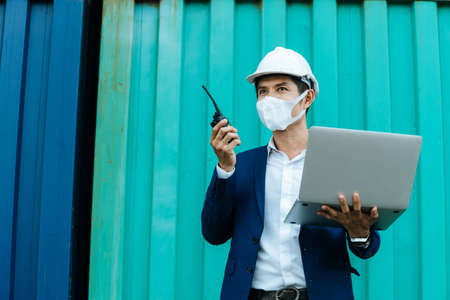 engineer control worker wearing protection face mask working on walkie talkie and digital laptop standing with container in background at cargo harbor, industrial, logistic import and export conceptの写真素材