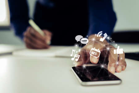 businessman hand using on digital mobile phone with virtual digital icon and writing on document report on desk at home office, digital marketing, social network, business finance, technology conceptの写真素材