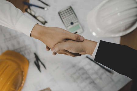 top view of architect, construction worker team hands shaking after plan project contract on workplace desk in meeting room office at construction site, contractor, partnership, construction conceptの写真素材