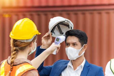 industrial worker woman temperature checking fever by digital thermometer with businessman with cargo container in background at container cargo harbor, industrial, logistic import and export conceptの写真素材