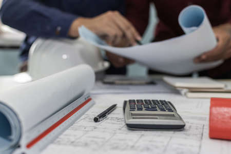 construction working tool, calculator, blueprint and safety helmet on architect workplace desk with architect in meeting room office center at construction site, construction, engineering tool conceptの写真素材