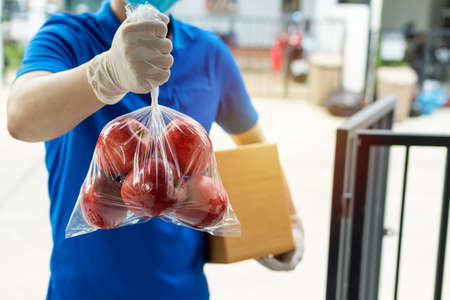 food delivery service man in blue uniform wearing protection face mask holding fresh red apple fruit waiting for customer at door home, express delivery, virus outbreak, takeaway food delivery conceptの写真素材