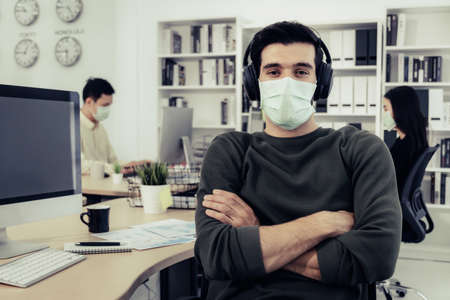 confident caucasian man in smart casual wearing protective face mask arms crossed and sitting in modern office with colleagues in background, social distance, new normal, business startup conceptの写真素材