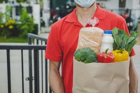 smart delivery man with protection face mask in red uniform holding fresh food set bag waiting for customer at door home, healthy food, express delivery, food delivery, online shopping conceptの写真素材