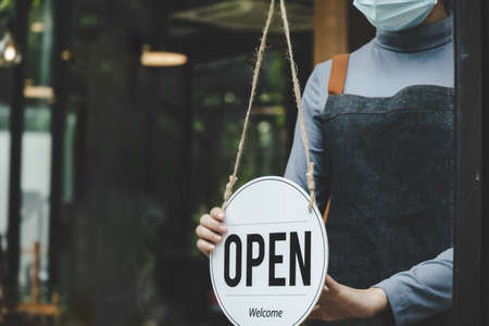 Reopen. friendly waitress wearing protection face mask turning open sign board on glass door in modern cafe coffee shop, cafe restaurant, retail store, small business owner, food and drink conceptの写真素材
