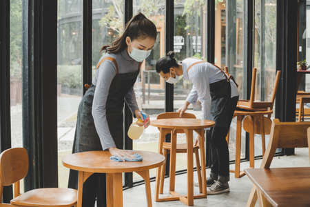asian waitress staff wearing protection face mask in apron cleaning table with disinfectant spray for protect infection coronavirus (covid-19) in cafe coffee shop restaurant. new normal conceptの写真素材