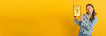 Valentines day. wide banner. happy asian pretty woman in casual clothing jeans smiling and hold new year gift box over copy space yellow color background, christmas celebration, happy hew year conceptの写真素材