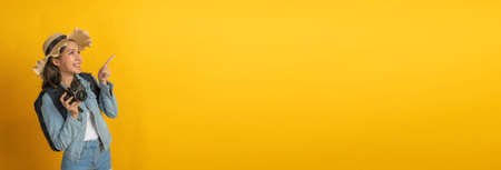 Backpacker. wide banner. asian woman hipster traveler with camera and hat in casual clothing happy and smiling over copy space yellow color background, summer holiday, backpacker travel conceptの写真素材