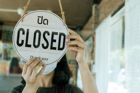 Lockdown. woman staff wearing protection face mask turning CLOSED sign board with Thai language on glass door in cafe coffee shop, cafe restaurant, small business owner, food and drink conceptの写真素材