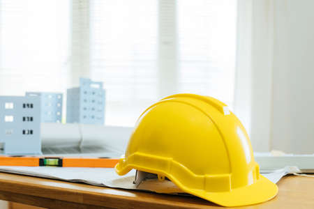 yellow safety helmet on workplace desk with building model, blueprint and tools in office center at construction site, partnership, industrial, construction contract and contractor conceptの写真素材