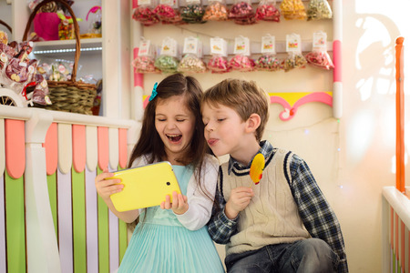 Happy kids looking at the tablet. Boy shows tongue holding candy in the handsの写真素材