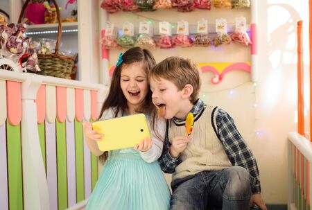 Happy kids looking at the tablet. Boy shows tongue holding candy in the handsの写真素材