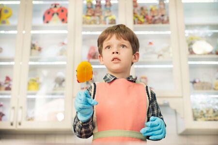 Little boy standing at kitchen in apron and holding lollipop in the handの写真素材