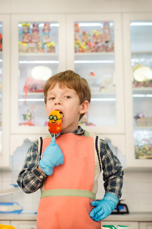 Little boy standing at kitchen in apron and licking candyの写真素材