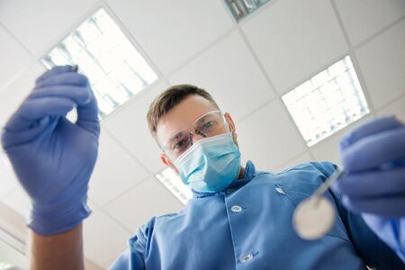 View from the patient side at the dentist. Young dentist in sterile mask and glasses approaching a patient with dental instruments in the handsの写真素材