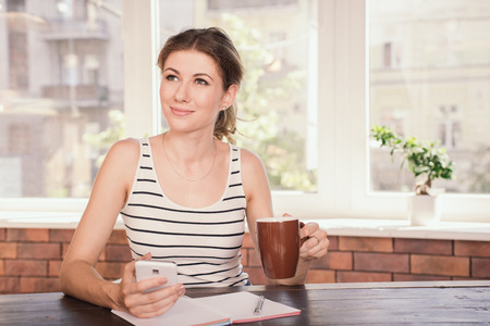Business woman working at home office. Young woman drinking coffee and typing message on her mobile phone while sitting at her desk.の写真素材