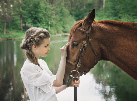 Woman standing with horse near lake in vintage dress and touching horse faceの写真素材