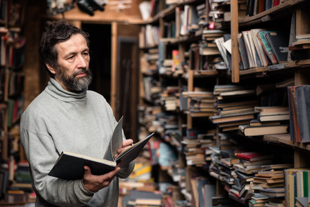 Portrait of authentic senior man with beard and good eyes reading book in the hand on  bookshelf backgroundの写真素材