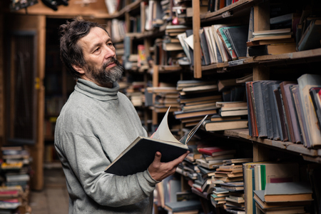 Portrait of authentic senior man with beard and good eyes reading book in the hand on  bookshelf backgroundの写真素材