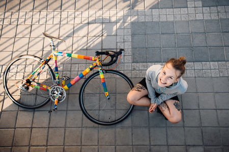 Happy young woman sitting near bicycle in the morning city at sunriseの写真素材