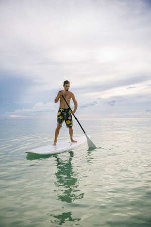 Young man stand up on paddle board and paddling out in tropical waters. Water sport near the beach...
