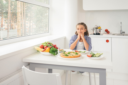 Cute little girl sitting by table in the kitchen and ready to have meal. Concept healthy lifestyle in modern familyの写真素材