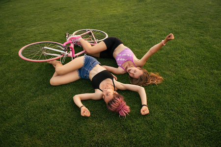 Joyful girls lying on the green grass with bicycle. Outdoor shot of happy two girls on bicycle. Top view.の写真素材