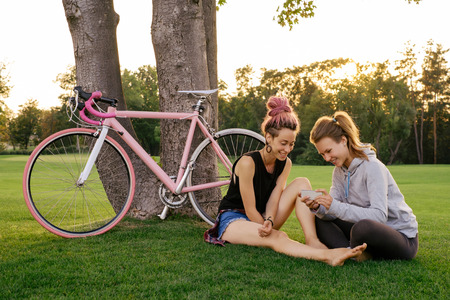 Two hipster woman with bicycle in city park having fun together. Outdoor shot of happy young female friendsの写真素材
