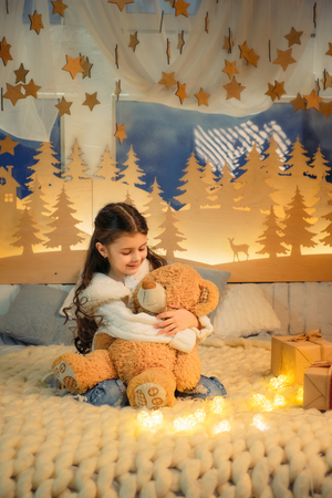 Cute little girl playing with teddy bear bed in room decorated for New Year. Winter weekends. Cozy scene. Holiday atmosphere.の写真素材