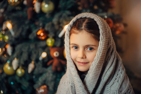 Cute little girl sitting near christmas tree in the evening and covered in warm scarf waiting for holiday. Cold winter weekends. Cozy sceneの写真素材