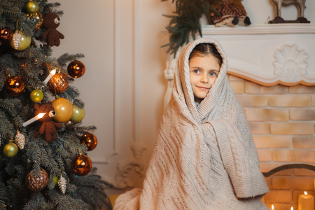 Dreamy little girl sitting near christmas tree in the evening and covered in warm scarf waiting for holiday. Cold winter weekends. Cozy sceneの写真素材