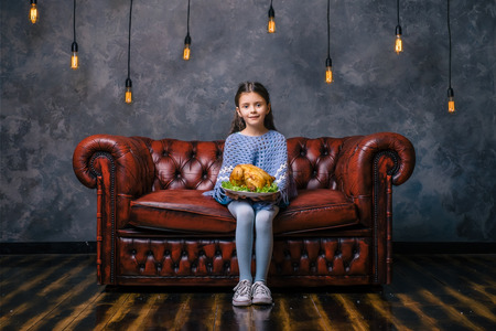 Little hungry girl sit on the sofa at night time and holding plate with tasty fried chicken. Taste food concept. Thanksgiving holiday.の写真素材