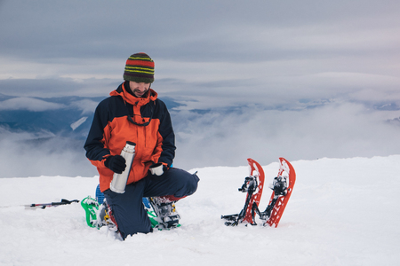 Man with backpack and snowshoes hiking in winter mountain and stopped to have rest and drink hot tea from thermosの写真素材
