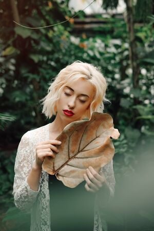 Fashion portrait of beautiful young with blond hair and perfect figure posing in the palm leaves in the jungle. Young pretty girl in green tropical forest or greenhouseの写真素材