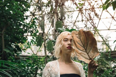 Beautiful young fashion woman with blond hair and perfect figure posing in the palm leaves in the jungle. Young pretty girl in green tropical forest or greenhouseの写真素材