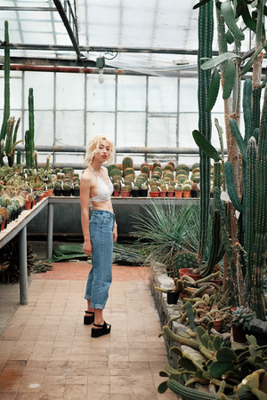 Fashionable portrait of beautiful young with blonde hair and red lips posing with perfect figure in the succulent cactus garden in the tropical greenhouseの写真素材