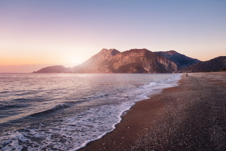 Woman enjoying beautiful sunrise on sea beach in Cirali village in Turkey. Rocky coastline, seashore with mountains on backgroundの写真素材
