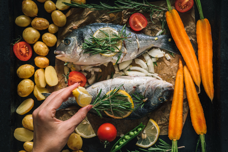 Preparing seafood fish for cooking on grill. Dorado fish with aromatic herbs and vegetables on rustic wooden board and dark background, top viewの写真素材