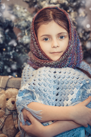 Cute little girl sitting near christmas tree in the evening and covered in warm scarf waiting for holiday. Cold winter weekends. Cozy sceneの写真素材