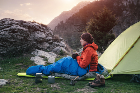 Traveler sitting on the ground next to tent holding cup with coffee in her handsの写真素材