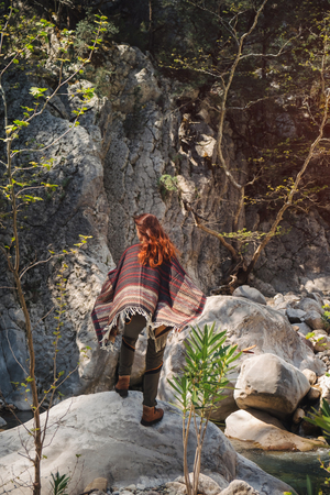 Red-head tourist in scattering poncho standing on the rock looking at the rivulet in the mountains on a sunny day. Shot from backの写真素材
