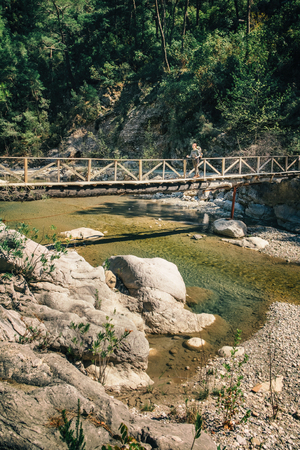 Distant silhouette of female traveler crossing the wooden bridge over the mountain rivulet on a sunny dayの写真素材
