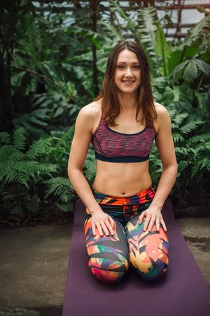 Attractive smiling female yoga instructor in a colorful sporty outfit sitting in a kneeling pose on a purple mat at botanical garden. Looking at camera. Healthy lifestyle concept.の写真素材