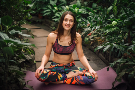 Beautiful barefooted female yoga instructor in a colorful outfit sitting in a easy pose, relaxing on a mat at greenhouse.Looking at camera. Healthy lifestyle concept.の写真素材