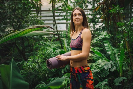 Smiling young caucasian woman in a sporty colorful clothes standing with yoga mat in her hands among palm leaves at greenhouse. Low angle view. Healthy lifestyle concept.の写真素材