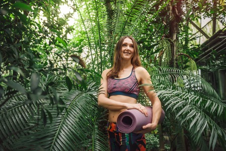 Smiling young caucasian woman in a sporty colorful clothes standing with yoga mat in her hands among palm leaves at greenhouse. Low angle view. Healthy lifestyle concept.の写真素材