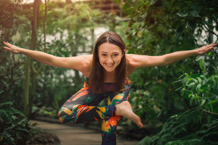 Portrait of smiling barefooted caucasian female yoga instructor in colorful sporty outfit performing standing pigeon pose on a sunny day at botanical garden. healthy lifestyle concept.の写真素材