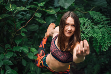 Barefooted sporty young caucasian female yoga instructor in a colorful outfit standing in a bow pose, stretching, doing mudra in forest. Healthy lifestyle concept. High angle view.の写真素材