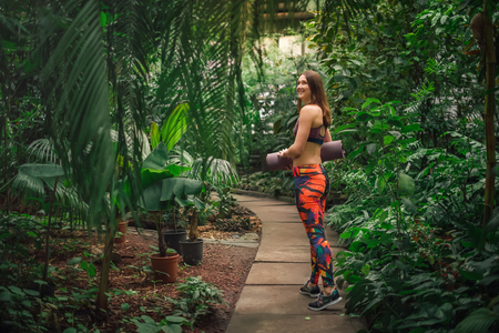 Full length portrait of beautiful smiling dreamy young woman in a colorful sporty outfit standing on a path, holding a mat at the botanical garden.Healthy lifestyle concept.の写真素材
