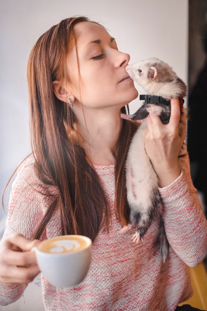 Sensual girl with a cup of cappuccino holding her pet ferret close to her lips in cafe. Bonding moment. Woman and a pet concept.の写真素材